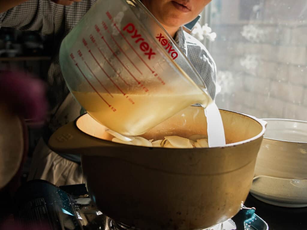 A person pouring liquid from a Pyrex measuring cup into a pot on a stove. The bright natural light from a window illuminates the scene, highlighting the kitchen setting.